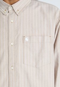Beige skjorte med knapper og hvide lodrette striber, med en lomme på venstre bryst og let, struktureret stof.