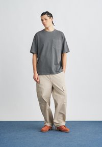 Szürke, túlméretezett póló mellzsebbel, bézs cargo nadrággal, melyben oldalzsebek találhatók. Narancssárga cipők texturált talppal, kék háttéren.