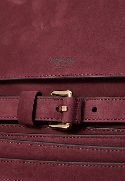 Burgundijas zamša soma ar izšūtām horizontālām līnijām, zelta sprādze un "MOSCHINO Milano" iedobījums virs sprādzes.