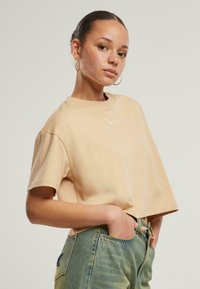 Beige lyhyt cropped-t-paita pehmeästä kankaasta, jossa on lyhyet hihat ja hienovarainen valkoinen brodeerattu logo rinnassa. Yhdistetty korkeavyötäröisiin farkkuihin.