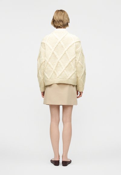 Άτομο που στέκεται και κοιτάζει προς τα πίσω, φοράει μια κρεμ παχιά πλεκτή μπλούζα, μπεζ φούστα και σκούρα επίπεδα παπούτσια σε απλό φόντο.