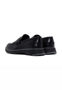 Zapatos negros de piel de tipo slip-on con una suela baja y texturizada. Presentan un acabado suave y un sutil detalle de costura en la parte superior y el talón.