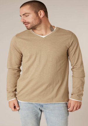 Man die een beige shirt met lange mouwen en V-hals draagt over een wit ondershirt en lichtblauwe spijkerbroek, kijkend naar links tegen een effen achtergrond.