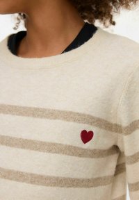 Pull beige avec des rayures horizontales beige, col rond, et un petit cœur rouge brodé sur le côté gauche. Texture douce, coupe décontractée.
