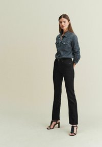Donkerdenim overhemd met knopen en voorkruiszakken, gedragen met strak passende zwarte jeans en zwarte haksandalen. Eenvoudig, strak design.