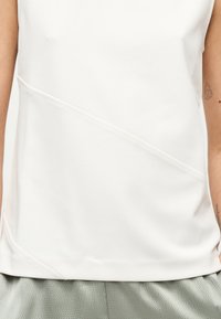 Λευκή αμάνικη μπλούζα με διαγώνια ραφή που φοριέται από άτομο με τατουάζ και στα δύο χέρια, σε συνδυασμό με ανοιχτό γκρι υφασμάτινο παντελόνι.