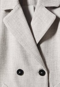 Cappotto doppiopetto grigio chiaro con un tessuto testurizzato, dotato di bottoni neri prominenti e rever affilati.