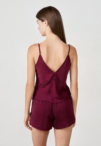 Kvinna med långt brunt hår som bär en djup röd satängcamisole och matchande shorts, visad bakifrån mot en ljus bakgrund.