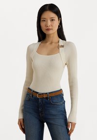 Vrouw met lang zwart haar, gekleed in een crèmekleurige ribbelige lange mouwen top met gespdetails en hooggewende blauwe jeans met een bruine riem.
