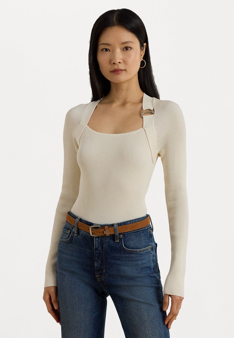Vrouw met lang zwart haar, gekleed in een crèmekleurige ribbelige lange mouwen top met gespdetails en hooggewende blauwe jeans met een bruine riem.