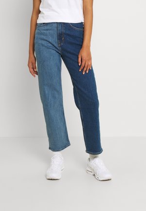 Persoon draagt een high-waisted, tweekleurige spijkerbroek met links een lichtblauwe en rechts een donkerblauwe pijp, gecombineerd met witte sneakers en sokken.