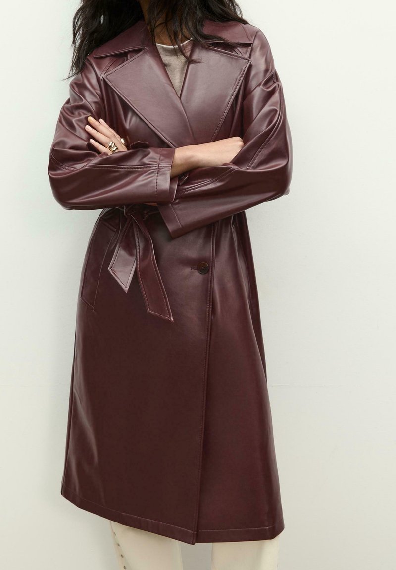 Femme portant un trench-coat en cuir marron avec une ceinture, debout les bras croisés devant un fond clair uni.