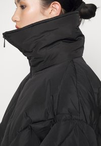 Chaqueta tipo puffer negra con un alto cuello, fabricada con un material suave y acolchado. Presenta puños elásticos y un diseño minimalista.