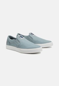 Zapatillas de lona azul sin cordones con una suela de goma blanca, que cuentan con una etiqueta de logo en la lengüeta y acentos laterales texturizados.