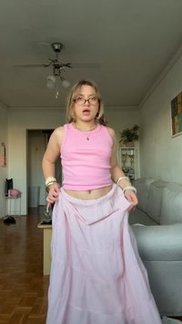 Jonge vrouw in een roze tanktop en rok terwijl ze haar rok aanpast in de woonkamer met een bank, planten, trofeeën en een plafondventilator.