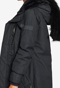 Abrigo de invierno negro con bolsillo con solapa abotonada en la manga, cierre frontal con cremallera y gran capucha forrada de piel, llevado por una persona de perfil lateral.