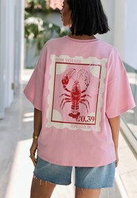 Lyserød oversized T-shirt med en hummergrafik i rødt, indrammet af et hvidt stempel med teksten "BON VOYAGE" og en pris på €0,39.