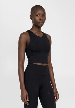 Atletisk kvinde med mørk hud og barberet hoved iført sort ærmeløs crop top og højtaljede leggings, stående mod en ensartet baggrund.