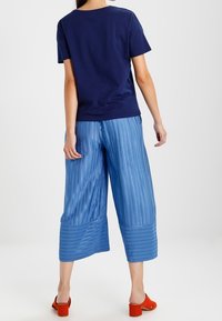 Žena v námořnické modré blůze s krátkým rukávem, světle modrých širokých kalhotách s pruhy a oranžových sandálech na podpatku, stojí zády k fotoaparátu.