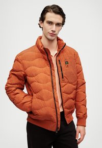 Narančasta prešana puffer jakna s patentnim zatvaračem, stojećim ovratnikom i rebrastim manžetama. Ima mali džep na prsima i toniranu mrežastu šaru.