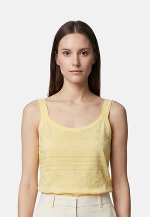 Jeune femme aux longs cheveux bruns et raides porte un haut sans manches brodé jaune pâle et un pantalon beige clair sur un fond uni.