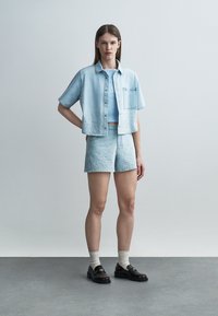 Jauna sieviete stāv pret vienkāršu fonu, valkājot gaisētas zilas džinsa šortus un īsas piedurkņu kreklu ar pogām, baltas zeķes un melnas loafers kurpes.