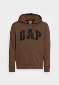 Sudadera con cremallera marrón, hecha de tejido suave, que presenta un gran logo negro de "GAP" en el pecho, bolsillos frontales y capucha ajustable.