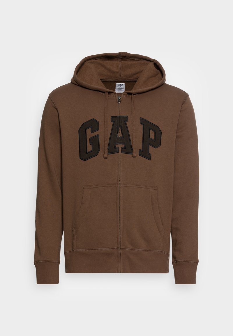 Sudadera con cremallera marrón, hecha de tejido suave, que presenta un gran logo negro de "GAP" en el pecho, bolsillos frontales y capucha ajustable.