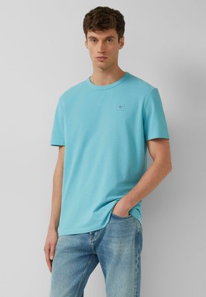 Junger Mann, der ein hellblaues T-Shirt mit Rundhalsausschnitt und hellblaue Jeans trägt, steht mit einer Hand in der Tasche vor einem einfarbigen Hintergrund.