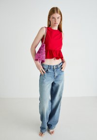 Kvinne i rød ermeløs crop topp, lyseblå løse jeans, hvite sandaler og store hoops øredobber, som bærer en rosa frynse håndveske over skulderen.