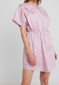 Vestito rosa con bottoni, caratterizzato da un ampio colletto ondulato, maniche corte e una vita elasticizzata per una vestibilità comoda. Tessuto dalla superficie liscia.