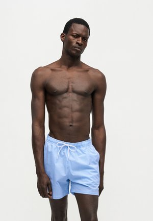 Homme à la peau foncée avec une musculature développée portant un short de bain rayé bleu clair et blanc sur un fond blanc uni.