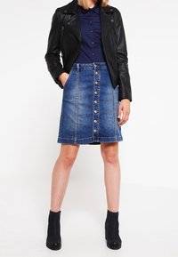 Falda de denim con corte recto, de color azul claro, cierre con botones en la parte delantera y dos bolsillos laterales, combinada con una chaqueta de cuero negra y botas de tobillo.