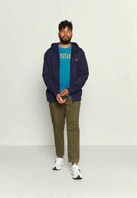 Felpa con zip blu marina sopra una maglietta grafica color teal, abbinata a pantaloni verde oliva e sneakers bianchi. Sovrapposizione casual con un design semplice.