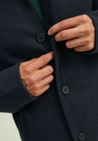 Cappotto di lana navy con grandi bottoni neri. Mani posizionate vicino ai bottoni, in evidenza la texture del tessuto e una fodera verde.
