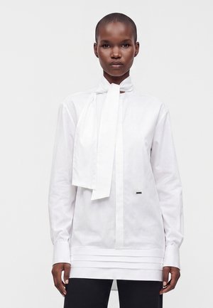 Personne portant un chemisier blanc à manches longues avec un grand détail de cravate et un pantalon noir, debout devant un fond clair uni.