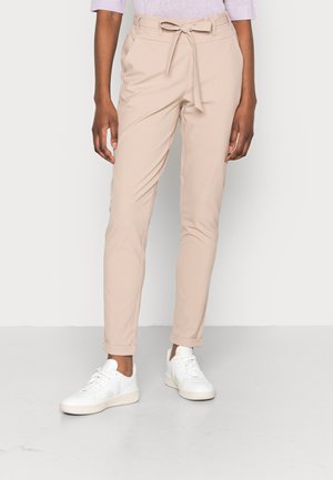 Personne portant un pantalon paperbag beige taille haute avec une ceinture nouée et des baskets blanches sur un fond clair uni.