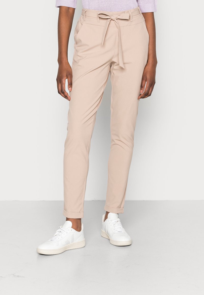 Personne portant un pantalon paperbag beige taille haute avec une ceinture nouée et des baskets blanches sur un fond clair uni.