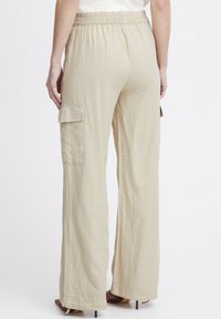 Pantalon large beige avec une taille élastique, deux poches cargo sur les côtés et un tissu texturé. Ourlets ouverts avec une légère flamme.
