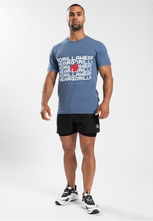 Blauwe katoenen t-shirt met vetgedrukte witte en rode tekst "GORILLA WEAR", gecombineerd met zwarte shorts en witte sneakers met zwarte accenten.