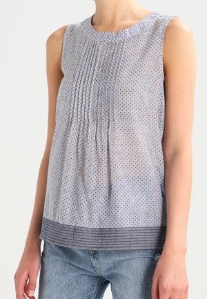 Mouwloze lichtgrijze blouse met verticale fijne strepen, geplooide voorkant en een donkerdere gestreepte zoom, gedragen met een blauwe spijkerbroek.