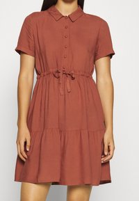 Robe à manches courtes, couleur rouille, avec une fermeture à boutons à l'avant, une taille à nouer et un ourlet à volants. Fabriquée dans un tissu doux et léger.