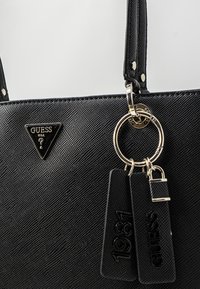 Sort tekstureret Guess håndtaske med guldfarvet beslag, trekantet logo og nøgleringsmærker, der læser "1981" og "GUESS" med en lille lås charm.