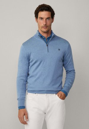Sweater zippé bleu clair avec une texture tricotée, doté d'un col montant et d'un logo brodé sur la poitrine, associé à une chemise bleue à col.