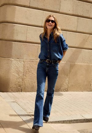 Denimhemd mit langen Ärmeln und Tasche, kombiniert mit blauem Schlagjeans. Dunkle Sonnenbrille und schwarze Stiefel. Glatte Textur, lässiger Stil.