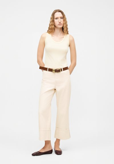 Γυναίκα με κυματιστά μαλλιά που φοράει μπεζ αμάνικη μπλούζα, μπεζ παντελόνι με φαρδιά πόδια και μανσέτες, καφέ ζώνη και σκούρα καφέ μπαλαρίνες, στέκεται με τα χέρια πίσω.