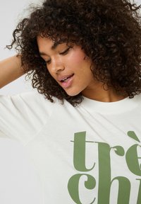 T-shirt blanc en matériau doux avec une coupe décontractée, arborant un design de texte vert "très chic" sur le devant. Manches courtes en raglan.