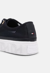 Černá teniska s texturovaným síťovaným svrškem, bílou platformovou podrážkou s geometrickým vzorem a malým logem na patě.