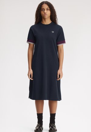 Donna vista frontalmente che indossa un vestito midi blu navy a maniche corte con bordi bordeaux e bianchi sulle maniche e scarpe nere con calzini.
