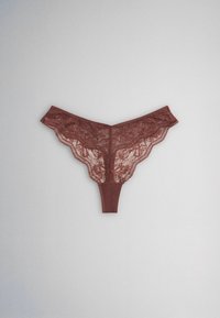 String en dentelle marron avec motif floral et bords festonnés sur fond gris clair.